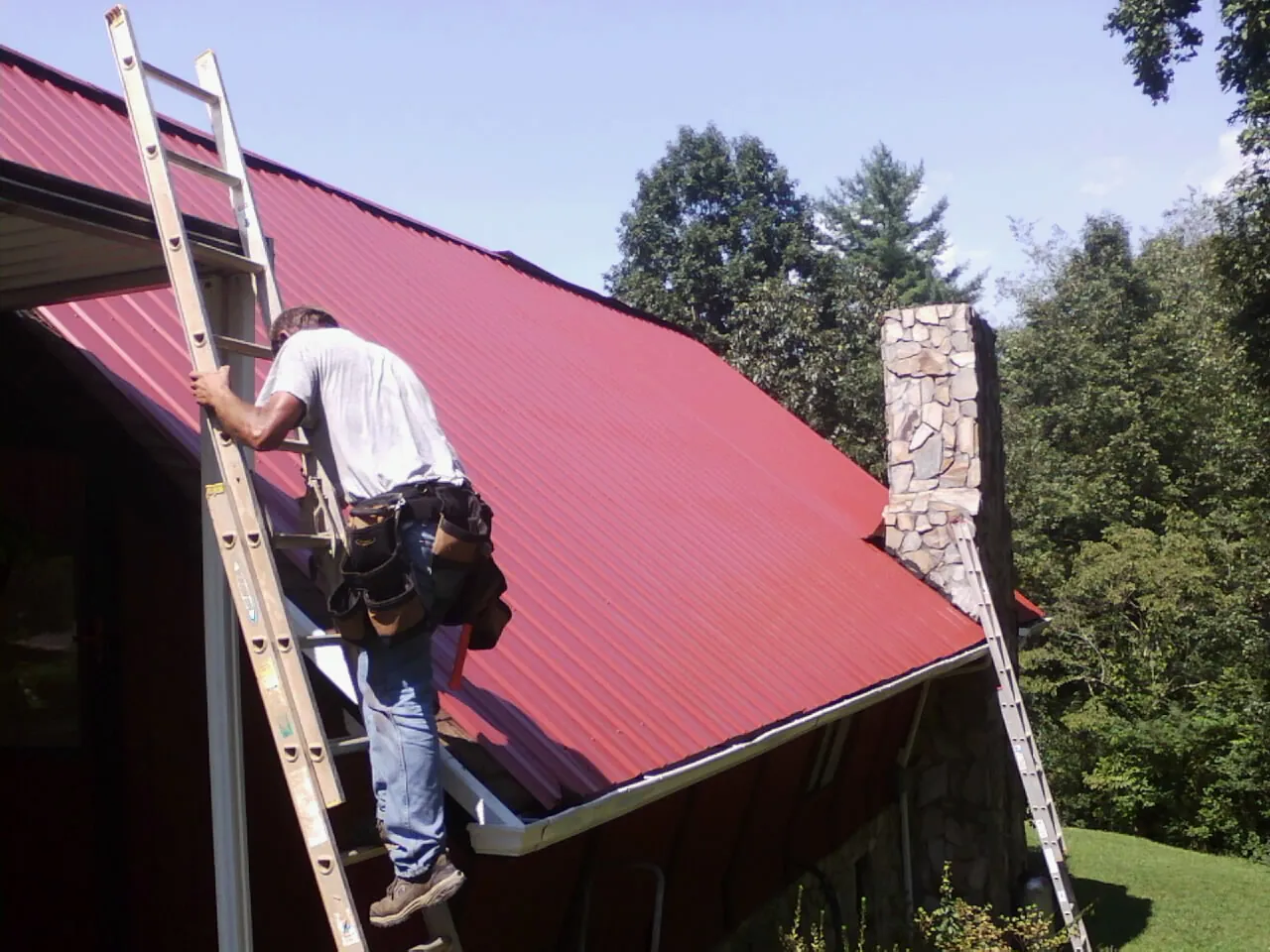 Roofer on a ladder beside a red metal roof and stone chimney — Soffit & Fascia Repair by Roofing Brothers in Rockaway Park